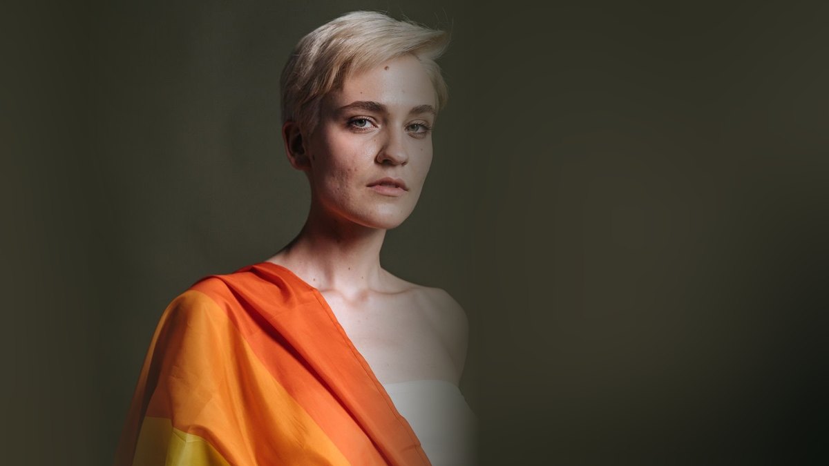 Confident person with a pride flag draped over the shoulder looking calmly at the camera in a respectful studio portrait